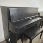 A black upright piano with a matching bench sits on a carpeted floor against a white wall.