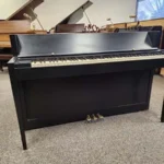 A black upright piano with worn keys stands on a carpeted floor, with grand pianos visible in the background.