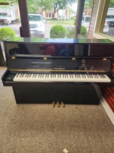 Black upright piano with three pedals, placed on a carpeted floor near large windows showing parked vehicles outside.