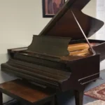 A brown grand piano with its lid open, accompanied by a wooden bench, sits against a beige wall under framed art.