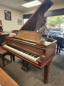 A brown grand piano with its lid open sits in a carpeted room with large windows and framed art on the walls.