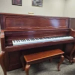Upright wooden piano with a matching bench in a carpeted room, artwork hanging on the wall in the background.