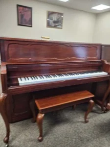 Upright wooden piano with a matching bench in a carpeted room, artwork hanging on the wall in the background.